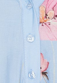 Lys blå bluse med knaplukning, blomsterprint detaljer i lyserød og blødt, struktureret materiale. Knappene er runde og klare.