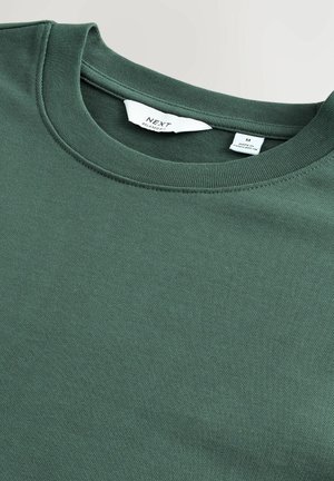 Hanorac verde, cu croi lejer, fabricat dintr-un amestec de bumbac, având guler rotund și o textură moale, netedă. Eticheta de îngrijire este vizibilă în interiorul gulerului.