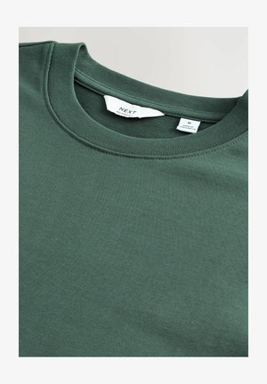 Hanorac verde, cu croi lejer, fabricat dintr-un amestec de bumbac, având guler rotund și o textură moale, netedă. Eticheta de îngrijire este vizibilă în interiorul gulerului.