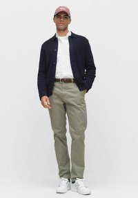 Herrklädsel med en mörkblå cardigan, vit skjorta, olivgröna byxor, brun flätad bälte och vita sneakers med turkosa detaljer.