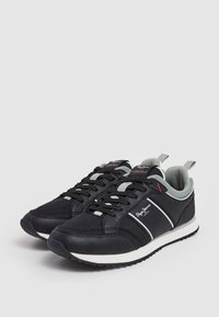 Paire de baskets basses noires et grises avec semelles blanches et panneaux latéraux marqués, exposée sur un fond clair uni.