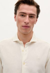 Beige button-up skjorte lavet af letvægtsstof, med en klassisk krave, subtile syninger og et lille broderet logo på brystet.