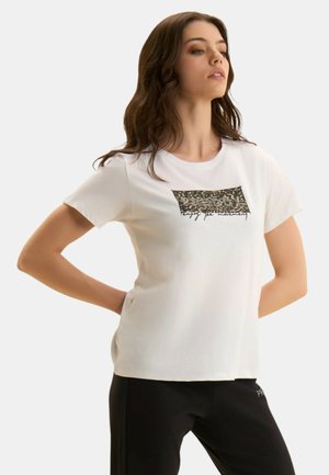 Giovane donna con lunghi capelli castani che indossa una maglietta bianca a maniche corte con stampa leopardata e la scritta "enjoy the movement", posa con le mani dietro la schiena.