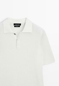 Maglietta polo bianca in cotone con trama a tessuto, maniche corte e colletto classico; presenta un unico bottone sopra la patta.