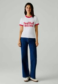 Mulher de pé, vestindo uma t-shirt branca da Red Bull Racing, jeans azuis e ténis brancos, em frente a um fundo cinza simples.