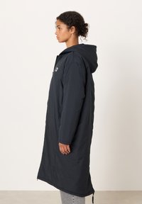 Kvinna stående i profil, iklädd en svart lång kappa med huva och dragkedja samt ljusgrå byxor, mot en enfärgad ljus bakgrund.