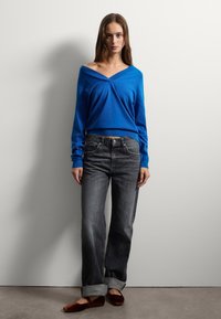 Pull bleu à épaules dénudées avec une coupe ample, associé à un jean gris foncé légèrement évasé et des chaussures plates marron, se tenant devant un mur uni.