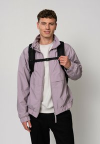 Giacca viola chiaro con zip, colletto rialzato, polsini elasticizzati e due tasche frontali, indossata sopra una camicia bianca e abbinata a uno zaino nero.