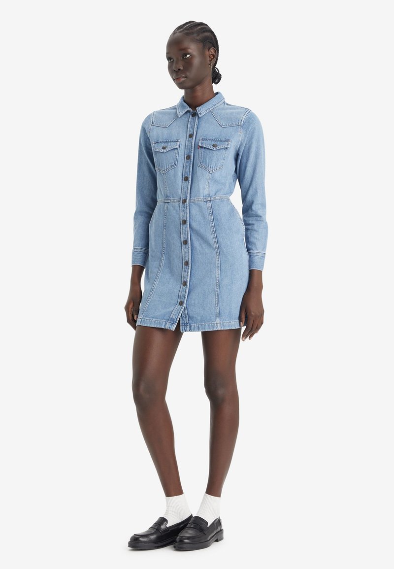 Robe chemise en denim bleu clair, avec un devant à boutons, des manches longues, deux poches poitrine et une silhouette ajustée. Col plat et détails cousus.