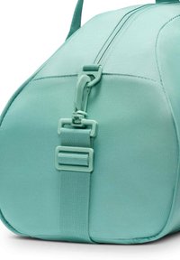 Nærbilde av en turkis stoff duffelbag som viser side stropp med plastspenne og glidelåsdetalj mot en hvit bakgrunn.