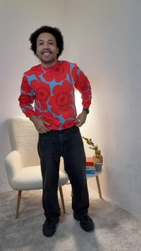 Man met krullend haar die lacht, gekleed in een rood-blauw bloementrui, zwarte broek, staand naast een crèmekleurige stoel en een klein tafeltje met boeken en een vaas.
