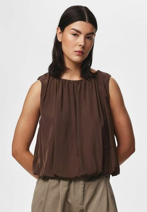 Femme aux cheveux foncés portant un top marron ample sans manches et un pantalon beige, debout devant un fond clair uni.