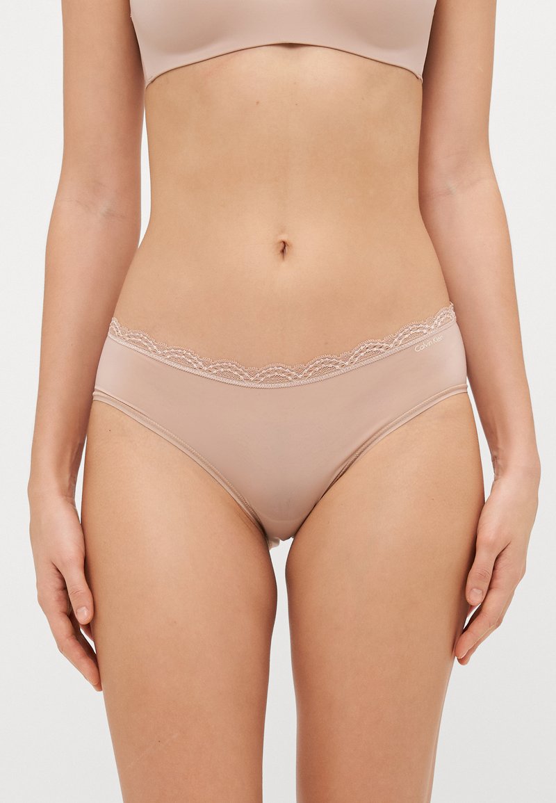 Beige truse med blonder kanter og glatt tekstur, med en bølget midje og en tettsittende design. Materiale i bomullsblanding for komfort.