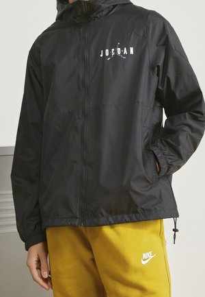 Persona con una chaqueta Jordan negra con cremallera y pantalones deportivos amarillos Nike, de pie con una mano en el bolsillo contra un fondo liso.