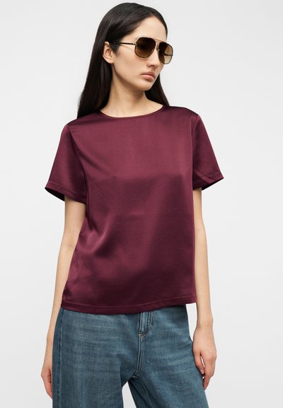 Μπορντό τοπ με κοντά μανίκια, λεία υφή και στρογγυλεμένο λαιμόκοψη, συνδυασμένο με φαρδιά μπλε τζιν παντελόνια. Το μοντέλο φοράει υπερμεγέθη γυαλιά ηλίου.