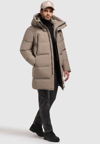 Jachetă puffer bej cu glugă, având buzunare cu fermoar și o textură netedă. Asortată cu blugi negri și cizme negre.