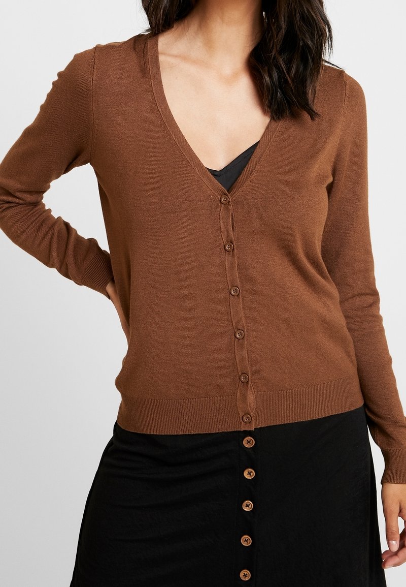 Femme portant un cardigan marron boutonné sur un haut noir et une jupe noire avec des boutons à l'avant.