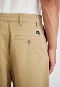 Khaki trousers with a straight fit, featuring a back pocket with a button closure and a branded patch. Cotton material, smooth texture.