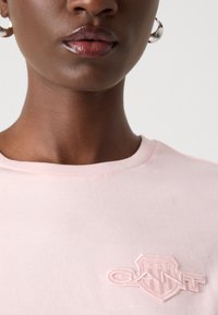 Helles rosa Baumwoll-T-Shirt mit einem erhabenen, gestickten Logo auf der linken Brust, einem Rundhalsausschnitt und einer glatten Textur.