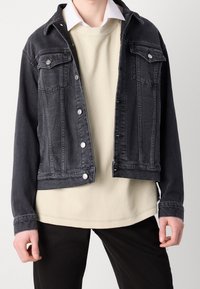 Svart denimjacka med knappstängning, två bröstfickor och en avslappnad passform, som är lager-på-lager över en ljusbeige strukturerad skjorta.