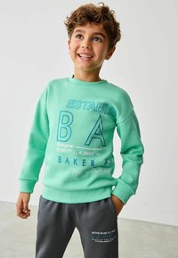 Mintgrön sweatshirt med grafisk text i mörkblått, rund halsringning och ribbade ärmslut. Tillsammans med grå sweatpants med en logo.