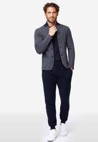 Blazer in maglina blu navy con accenti grigi, monopetto, due tasche; abbinato a jogger scuri e sneakers bianche, creando un ensemble casual.