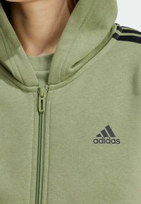 Sudadera con capucha y cremallera en verde claro, hecha de tejido suave, con rayas negras como acento y un logotipo de Adidas bordado en el pecho.