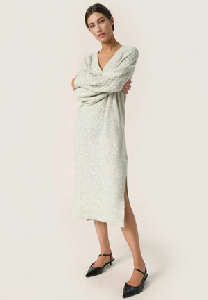 Langes, hellgraues gestricktes Kleid mit V-Ausschnitt, strukturiertem Muster und seitlichem Schlitz. Tragen mit schwarzen, spitzen Schuhen und Knöchelriemen.