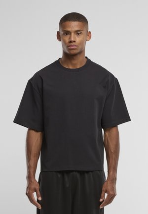 Nuori mies, jolla on lyhyet hiukset, pukeutuneena väljään mustaan lyhythihaiseen t-paitaan ja mustiin housuihin, seisoo yksivärisen vaalean taustan edessä.