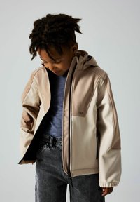 Leichte Kapuzenjacke in Beige und Creme mit strukturierten Einsätzen, Reißverschluss und verstellbaren Bündchen. Getragen über einem blauen Hemd und dunkler Jeans.
