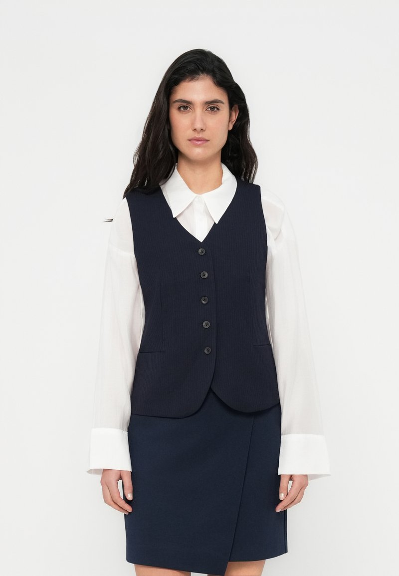 Γιλέκο ναυτικού μπλε με ριμπ υφή, πέντε κουμπιά κατά μήκος του μπροστινού μέρους και δύο εμπρόσθιες τσέπες, σε συνδυασμό με λευκό πουκάμισο με γιακά και φούστα ναυτικού μπλε.