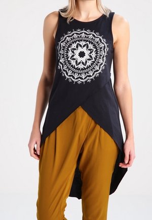 Femme portant un haut noir sans manches avec un motif mandala blanc et un ourlet asymétrique, associé à un pantalon jaune moutarde.
