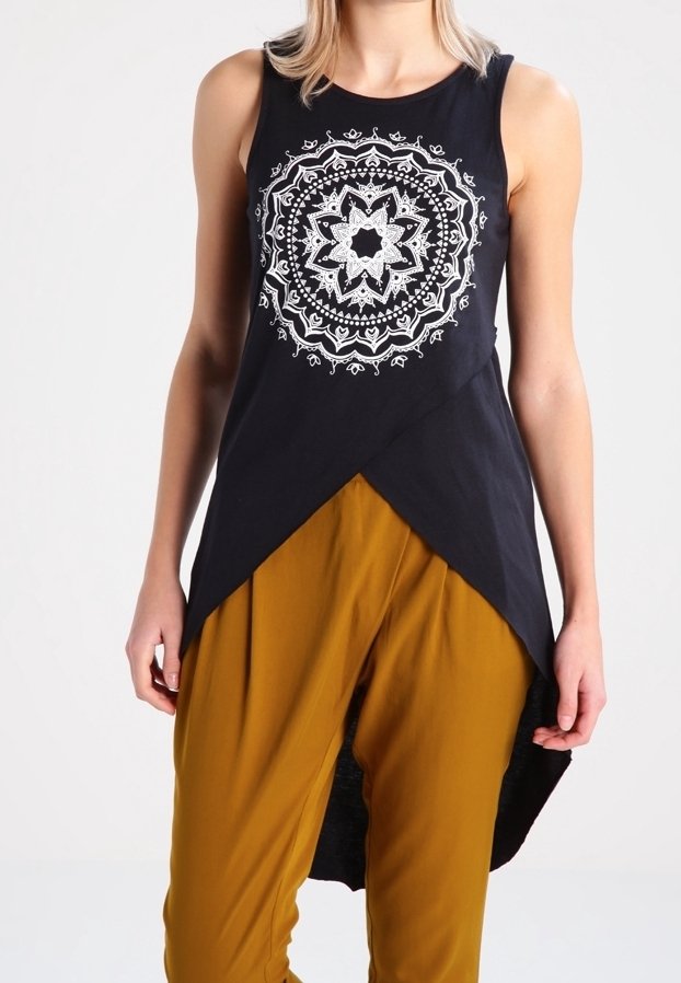 Femme portant un haut noir sans manches avec un motif mandala blanc et un ourlet asymétrique, associé à un pantalon jaune moutarde.