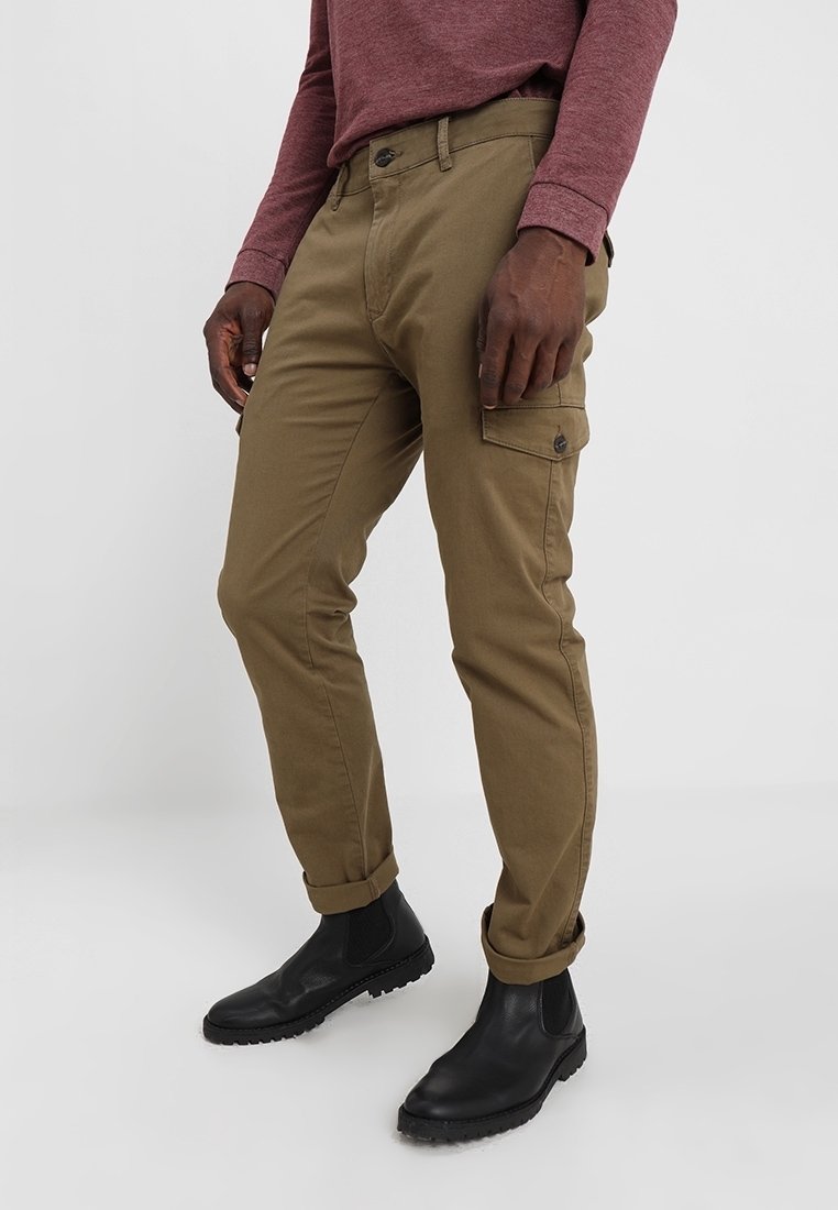 Muž v olivově zelených cargo kalhotách s ohrnutými nohavicemi, černých kožených botách a vínovém tričku s dlouhým rukávem, stojící na neutrálním pozadí.