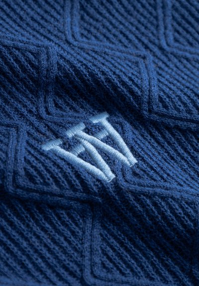 Tissu tricoté bleu avec un motif texturé de lignes verticales et diagonales. Des initiales brodées en bleu clair mettent en valeur le design.