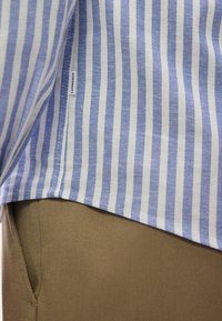 Μπλε και λευκό πουκάμισο με κάθετες ρίγες, φορεμένο μέσα από καφέ παντελόνι, με ετικέτα στην πλαϊνή ραφή που δείχνει την επωνυμία "LINDBERGH".