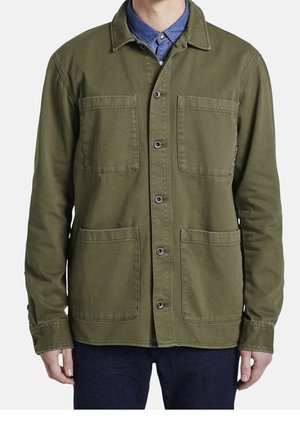 Mężczyzna noszący oliwkowozieloną kurtkę zapinaną na guziki z czterema przednimi naszywanymi kieszeniami, na niebieską koszulę z kołnierzykiem i ciemne spodnie.