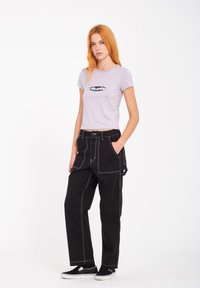 Fioletowy krótki t-shirt z logo oraz czarne spodnie o szerokich nogawkach z białymi strefami, kieszeniami bocznymi i pętlą na pasek. Czarne buty wsuwane.