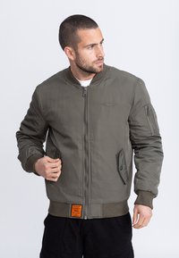 Hombre con cabello corto y barba, vestido con una cazadora bomber verde oliva con cremallera, puños y bajo acanalados, ajustando el bolsillo izquierdo, mirando hacia la derecha.