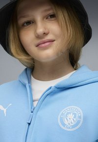 Modrá zipsová mikina vyrobená z mäkkého materiálu s prednou kapsou, na hrudi s logom Manchester City a bielym tričkom pod ňou.
