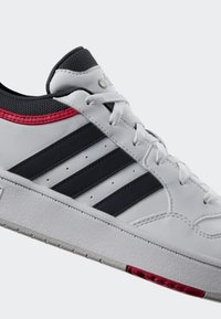 Scarpa sportiva bianca con strisce nere, colletto imbottito, accento rosso sul tallone e suola in gomma testurizzata per una presa migliore. Allacciata con lacci bianchi piatti.