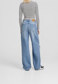 Donna che indossa un crop top a righe bianche e nere con maniche lunghe, jeans larghi di un blu chiaro e sneakers bianche e nere, in piedi con la schiena rivolta verso di noi.