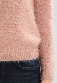 Rosa gestrickter Pullover mit flauschiger Textur, gerippten Bündchen und Saum, kombiniert mit dunkelblauer Jeans. Nahaufnahme, die die Stoffdetails hervorhebt.
