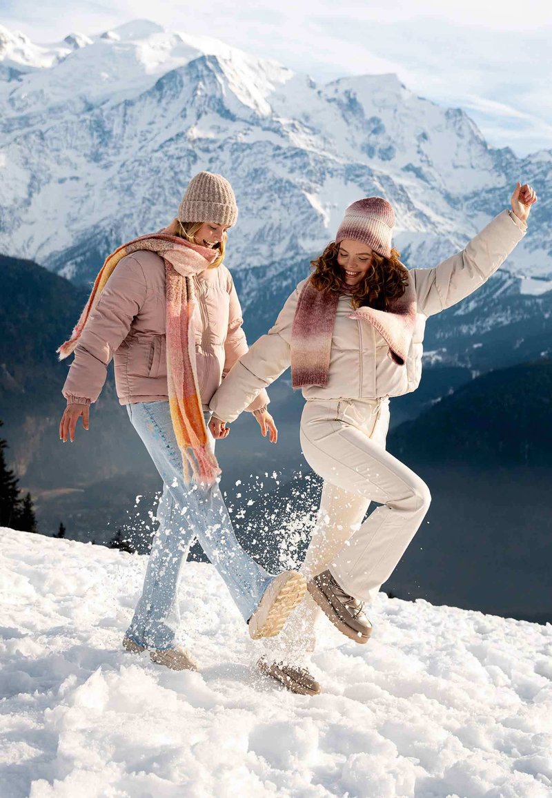 Twee mensen in winterkleding spelen in de sneeuw. De één draagt een roze pufferjack en een meerkleurige sjaal; de ander draagt een crèmekleurige jas en bijpassende broek.