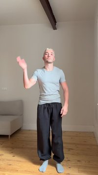 Jeune homme aux cheveux blonds courts, debout pieds nus sur un sol en bois, portant un t-shirt gris et un pantalon noir, levant la main gauche dans une pièce vide.