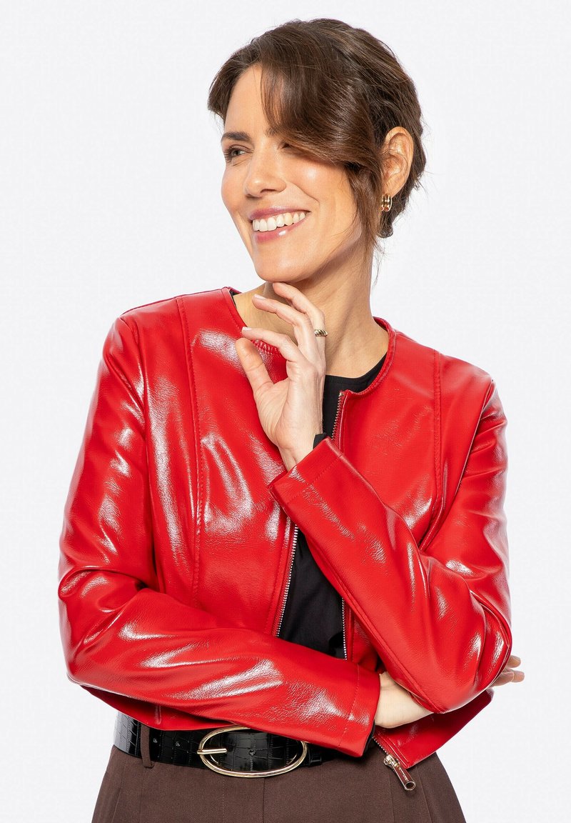 Veste en simili cuir rouge avec un col rond, fermeture éclair et manches longues. Texture brillante et coupe ajustée, associée à une ceinture noire.