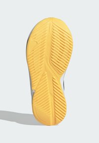 Suela de zapato con un destacado patrón de goma amarilla, que presenta surcos diagonales y verticales para tracción. Acentos en blanco y negro visibles.