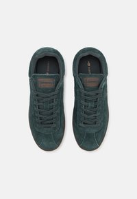 Zapatillas de ante verde con un acabado texturizado, con cordones planos, punta redonda y suela de goma marrón. Marca Lacoste en la lengüeta.
