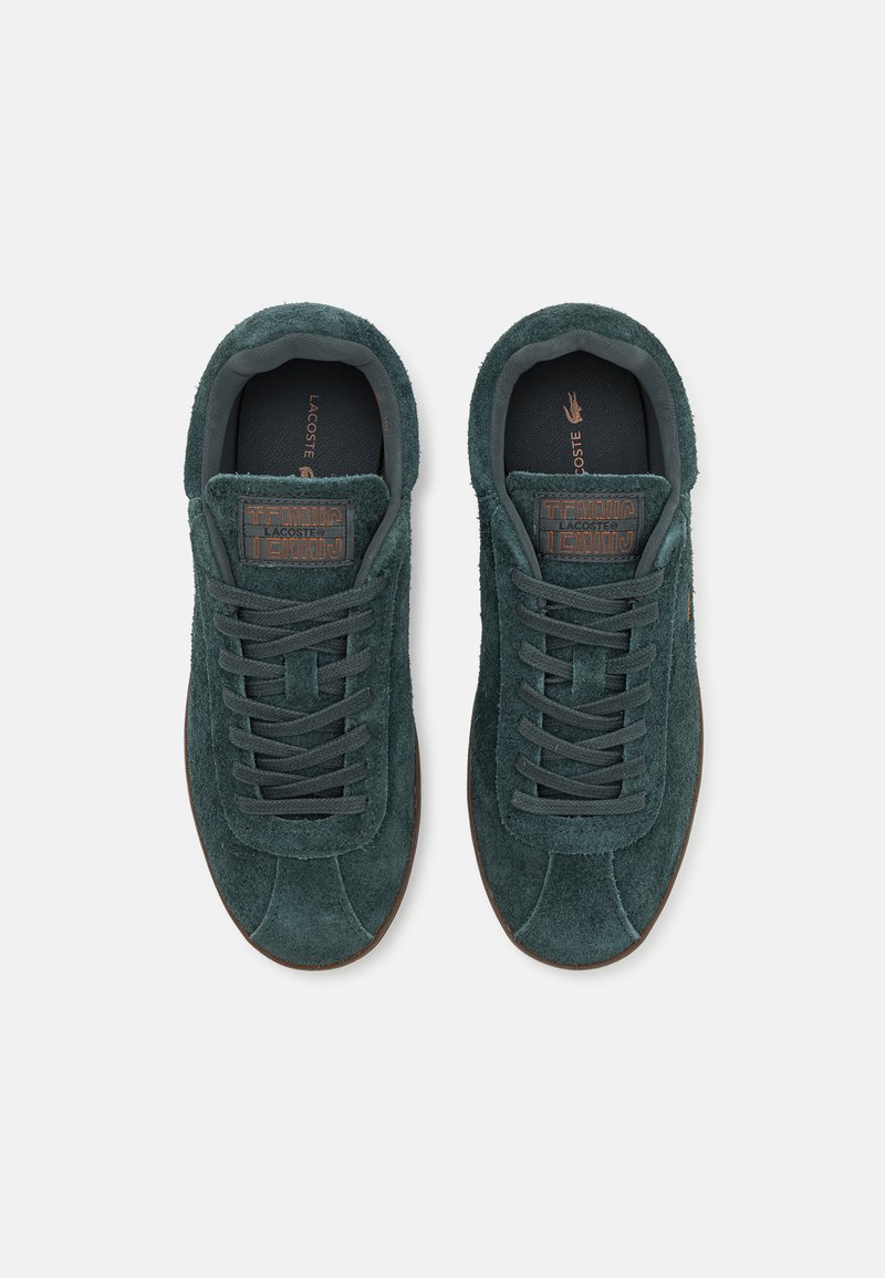 Zapatillas de ante verde con un acabado texturizado, con cordones planos, punta redonda y suela de goma marrón. Marca Lacoste en la lengüeta.