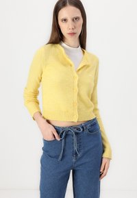 Cardigã amarelo com um corte cropped, punhos e bainha canelados, cinco botões e uma textura suave. Combinado com calças de ganga azul de cintura alta.